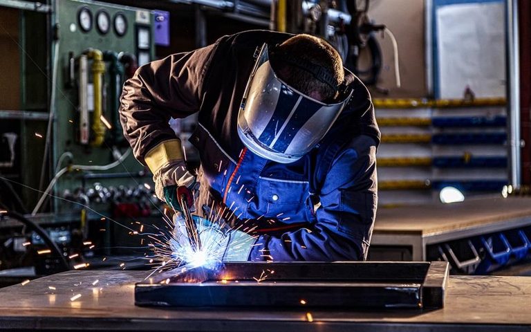 Welder in a full-face helmet and gloves welding a steel frame, sparks flying in a workshop